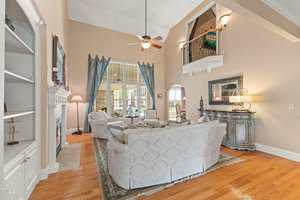 A spacious living room with high ceilings, featuring a matching set of patterned sofas and armchairs around a rug. There's a fireplace, built-in shelving, and large windows. A ceiling fan is overhead, and stairs lead to a small upstairs balcony.