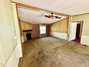 Interior of an empty room with a beige carpet, a ceiling fan, and a stone fireplace on the left. The walls are painted tan with striped wallpaper on one side. A window with blinds is centered on the far wall, and a doorway is visible on the right.