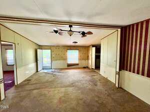 An empty living room with a beige carpet and cream walls, featuring a ceiling fan with lights. A striped red and cream accent wall is on the right. There are two windows with blinds and a door leading outside. A smaller room is visible on the left.