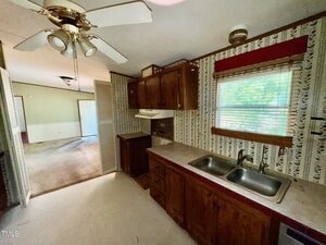 Kitchen with dark wood cabinets, stainless steel double sink, and floral-patterned wallpaper. Ceiling fan with lights above. Window with red valance. Open view to adjacent carpeted room.