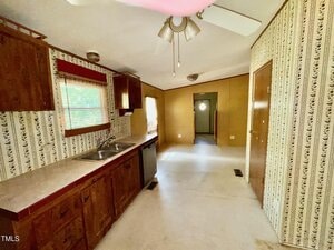 A kitchen with wooden cabinets, a white countertop with a sink, and patterned wallpaper. The ceiling has a fan and light fixture. There are windows on the left, and a corridor with doorways at the far end.