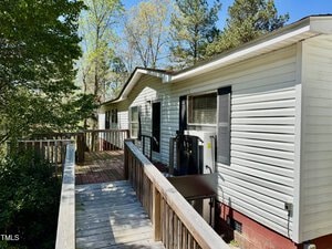 A white mobile home with a sloped wooden ramp leading to the entrance. The exterior features small windows with shutters and is surrounded by tall trees under a clear blue sky.