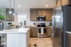 Modern kitchen with light wood cabinets, stainless steel appliances, and a white marble countertop. A potted plant and fruit stand adorn the island. Pendant lights hang above, and a window provides natural light. Light wood flooring completes the look.