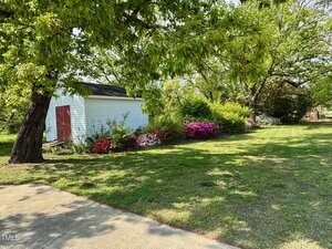A small white shed with a red door is surrounded by green grass and trees. Colorful flowers, including pink and purple blooms, line the edge of the building. The sky is clear, suggesting a sunny day. A concrete path is in the foreground.
