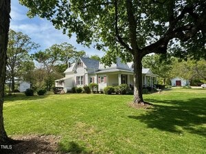 A white house with a gray roof sits on a spacious, grassy yard. Trees provide shade in the foreground, while a red outbuilding is visible in the background under a partly cloudy sky. Shrubs line the house's foundation.
