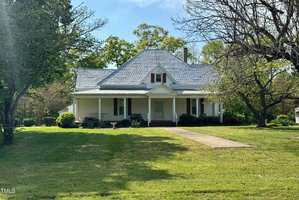 A quaint, single-story house with a light gray roof and beige siding sits amid a lush green lawn. A concrete pathway leads to the front porch, which is surrounded by trees and shrubs under a clear blue sky.