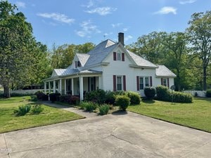 A white house with red shutters and a grey roof is surrounded by a green lawn and trees. There is a paved driveway in the foreground and a small garden with shrubs and flowers near the porch. The sky is clear and blue.
