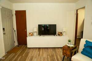 A living room with a flat-screen TV on a white console against a white wall. Two table lamps and decorative items are on the console. There is a wooden side table with a small plant next to a white armchair. The floor is light wood.