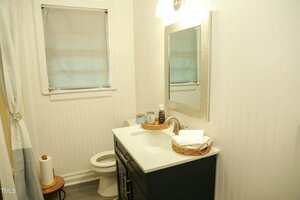 A small bathroom with white walls features a window with a gray shade, a vanity sink with a mirror above, and a toilet. A wicker basket with towels is on the sink, and a roll of toilet paper sits on a wooden stool beside the toilet.
