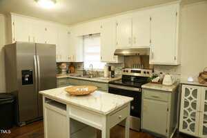 A modern kitchen at 513 Orange Street boasts white cabinets and a stainless steel refrigerator. The room features a small island with a marble countertop, an electric stove, and a double sink. A backsplash with a mosaic tile pattern adds detail to the decor.