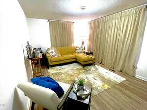 A living room with a yellow L-shaped sofa, a white armchair with a blue cushion, and a wooden floor. Curtains cover the windows, and a patterned rug lies on the floor. A lamp and small tables are placed around the seating area.