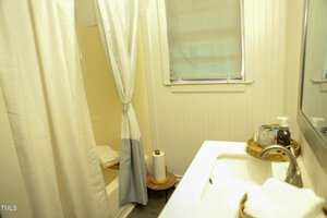 A small bathroom featuring a shower with a curtain, a window with a closed blind, a sink with a wicker basket holding a soap dispenser and a small bottle, a roll of paper towels on a stand, and folded towels on the bathtub edge.