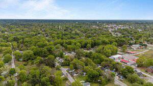 Aerial view of a small town with many trees, residential houses, and some commercial buildings. Roads intersect the area, and the landscape is mostly green with a partly cloudy sky above.