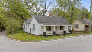 A small white house with a covered front porch and black trim sits on a corner lot with overgrown grass and yellow flowers. The house has a gabled roof and two chimneys, with trees and another house nearby.