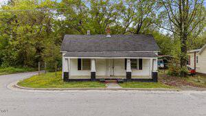 A small, single-story house with a covered front porch, white siding, black trim, and a dark shingled roof sits on curved 905 David Street in Henderson. Trees and greenery surround the home; another house is partially visible on the right.