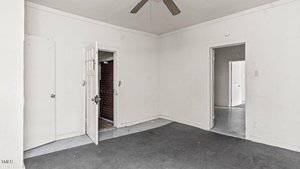 An empty, white-walled room with a dark carpet, ceiling fan, two open doors revealing other rooms, and one open door showing a brick wall and concrete floor. The room appears worn and has minimal decor.