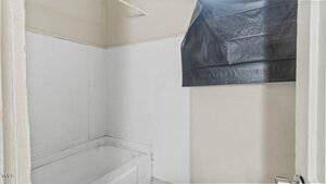 A small bathroom with a white bathtub, white walls, and a window covered by a black plastic sheet. The space appears empty and in need of renovation.