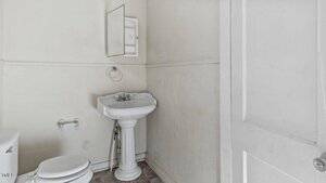 A small bathroom with a white pedestal sink, a wall-mounted mirror cabinet, a toilet, a towel ring, and a toilet paper holder. The walls are off-white and the floor has gray tiles.