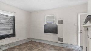 Empty room with white wooden walls and ceiling, two windows covered with black plastic, a wall heater between the windows, a door, and a floor with square brown tiles.