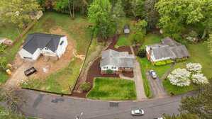 Aerial view of three houses along a curved residential street. One house has a dirt yard under construction, while the other two have landscaped yards with lawns, trees, and a small shed in the back. Two cars are parked on the street.