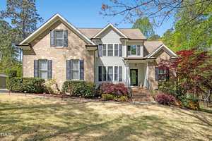 A two-story suburban house at 1503 Anterra Drive, Wake Forest, features a brick and light-colored siding exterior, black shutters, landscaped shrubs, and a front porch with steps, set on a sloped lawn with trees in the background.