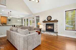 A living room with a large beige sectional sofa, a gas fireplace with a stone surround and wood mantel, a lamp, hardwood floors, and an open view into the kitchen and dining area.