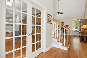 A hallway with hardwood floors features a staircase with wooden railings, a room with glass French doors on the left, and a ceiling fan with lights above. Furniture and wall art are visible in both rooms.