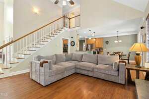 A spacious living room with a large gray sectional sofa on wooden floors, open to a kitchen and dining area with pendant lights and wooden cabinets, and a staircase leading to an upper level.