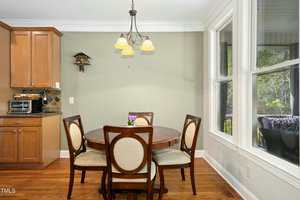 A small dining area with a round wooden table and four chairs sits next to large windows. A three-light chandelier hangs above the table, and wooden cabinets are visible on the left side of the image.