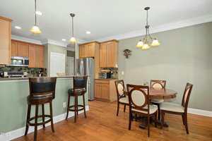 A kitchen and dining area with hardwood floors, light green walls, wooden cabinets, stainless steel appliances, a round dining table with four chairs, and a breakfast bar with three barstools.