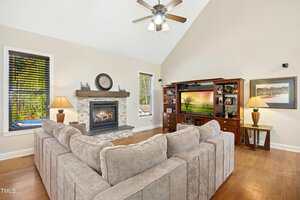 A living room with a beige sectional sofa, a stone fireplace with a wooden mantel, a ceiling fan, a large wooden entertainment center with a television, and two windows with blinds on either side of the fireplace.