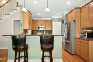 A kitchen with light wood cabinets, stainless steel appliances, a double-door refrigerator, granite countertops, two pendant lights, and two dark wooden barstools at the island. Stairs are visible in the background.