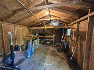 Interior of a wooden shed with exposed beams, workbench at the back, various tools, exercise equipment, plastic water jugs, and miscellaneous items scattered and stored along the walls and floor.