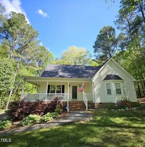 A single-story white house with a covered front porch, steps leading to the entrance, an American flag, and landscaped plants in the yard, surrounded by tall trees under a blue sky with some clouds.