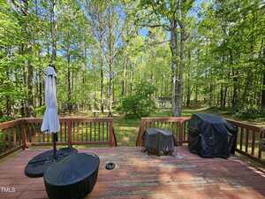 A wooden deck with two covered grills, a covered patio umbrella, and a small table overlooks a grassy backyard with trees and a small shed in the distance under a clear sky.