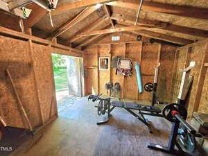 A home gym setup inside a wooden shed with exposed beams. There is a weight bench, weights, a barbell rack, and some equipment hanging on the wall. Sunlight comes through the open door.