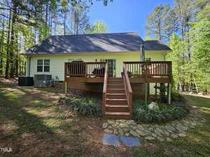 A white house with a dark roof and wooden deck, surrounded by trees. Stairs lead up to the deck with outdoor furniture and an umbrella. Stone steps and landscaping are visible in the foreground.