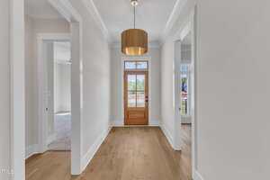 A hallway with light-colored walls and wood flooring leads to a wooden front door with glass panels. There are doorways to rooms on both sides and a wooden pendant light hanging from the ceiling.