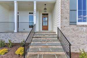 A stone walkway with black metal railings leads up to a wooden front door with glass panels. The entryway features white siding, brick accents, a hanging lantern, and the house number 3803. Shrubs are planted along the path.