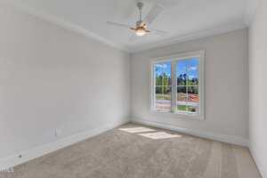 Empty room with beige carpet, white walls, a ceiling fan with light, crown molding, and a double window showing a view of trees and blue sky. Sunlight casts a shadow on the floor.