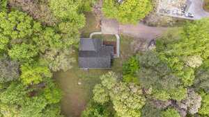 Aerial view of a house surrounded by green trees and lawn, with a driveway leading to a small parking area. A path curves from the driveway to the front entrance. A few vehicles are parked in the upper right corner.