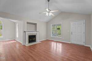 A living room with light beige walls, vaulted ceiling, ceiling fan with lights, white mantel fireplace, wood flooring, two large windows, and a white door; a bright adjacent room is visible through an open doorway.