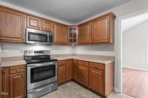 A kitchen with brown wooden cabinets, beige countertops, stainless steel oven and microwave, tiled floor, and an open doorway leading to a room with wood flooring.