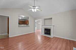 A living room with light-colored walls, wood-look flooring, a white ceiling fan, a fireplace with a white mantle, and open views into a kitchen and dining area through wide doorways.