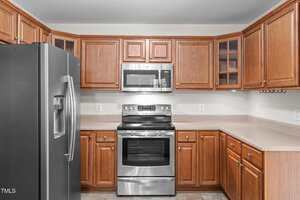 A kitchen with wooden cabinets, stainless steel refrigerator, electric stove, and microwave; beige countertops and tiled flooring are also visible.