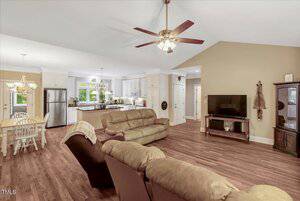 A living room with brown couches and a recliner faces a wall-mounted TV. The room has hardwood floors, ceiling fans, and an open view of a white kitchen and dining area with a table and chairs.
