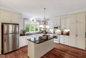 A modern kitchen with white cabinets, stainless steel refrigerator, central island with dark granite countertop, hardwood floor, pendant lights, and large window above the sink.