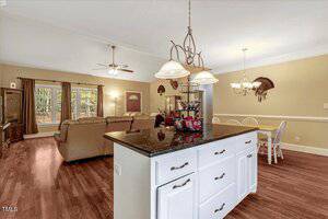 Open-concept living and dining area with light wood floors, beige walls, a white kitchen island with dark granite countertop, a dining table with four chairs, and a seating area with sofas and ceiling fans.