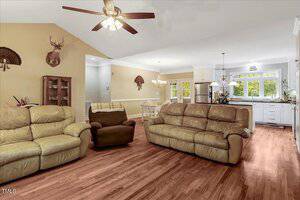 A living room with tan leather sofas, an armchair, hardwood floors, ceiling fan, and mounted deer decor on the wall. An open kitchen with white cabinets and dining area are visible in the background.