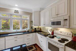 A modern kitchen with white cabinets, black granite countertops, a white electric stove with a microwave above it, a double sink beneath three windows, and small appliances on the counters. Natural light fills the room.